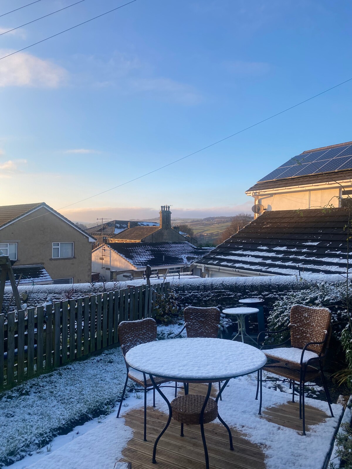 A winter scene captures a garden with a table and four chairs dusted in snow. The setting includes rooftops and hills in the background, with clear blue skies above and sunlight illuminating the landscape.