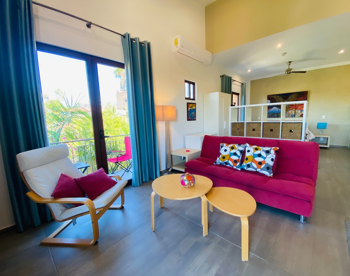 A bright living area features a vibrant red sofa adorned with decorative pillows. A natural wood coffee table complements a comfortable armchair. Large windows allow natural light to illuminate the space, with views of lush greenery visible outside.
