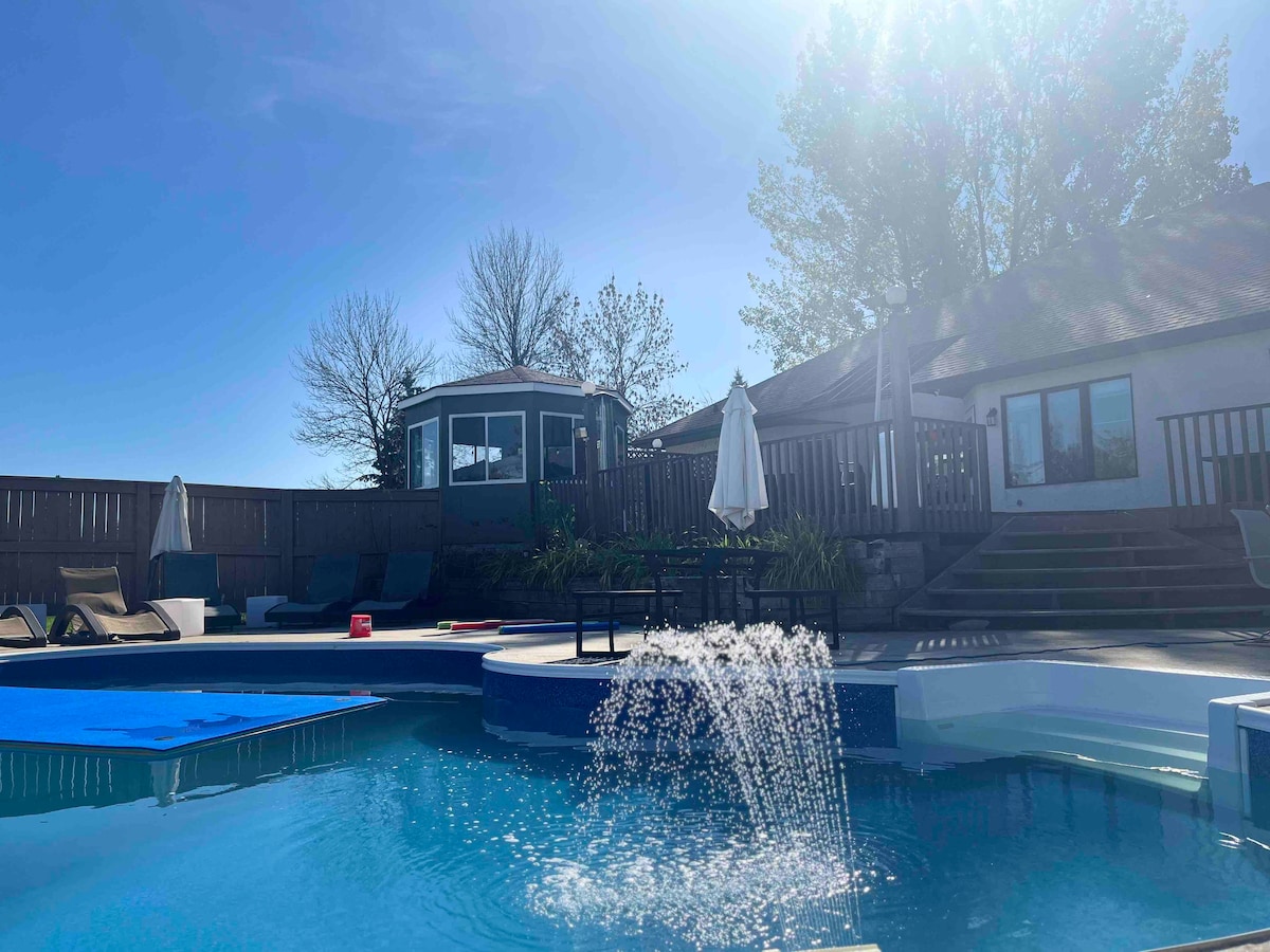 A sparkling pool is featured in the foreground with a fountain creating a gentle spray. Sunlight filters through the trees in the background, and a gazebo can be seen nearby. Umbrellas are positioned near lounge chairs along the pool's edge.