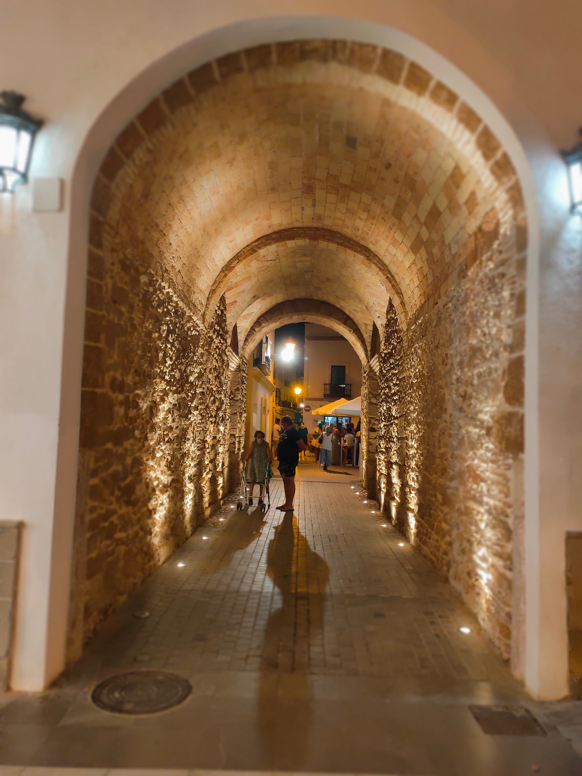 An arched passageway is lined with warm lighting that reflects off the textured stone walls. The cobblestone path leads to an inviting outdoor area, where small tables and umbrellas are visible in the distance. Two figures walk leisurely through the archway, creating a sense of tranquility.