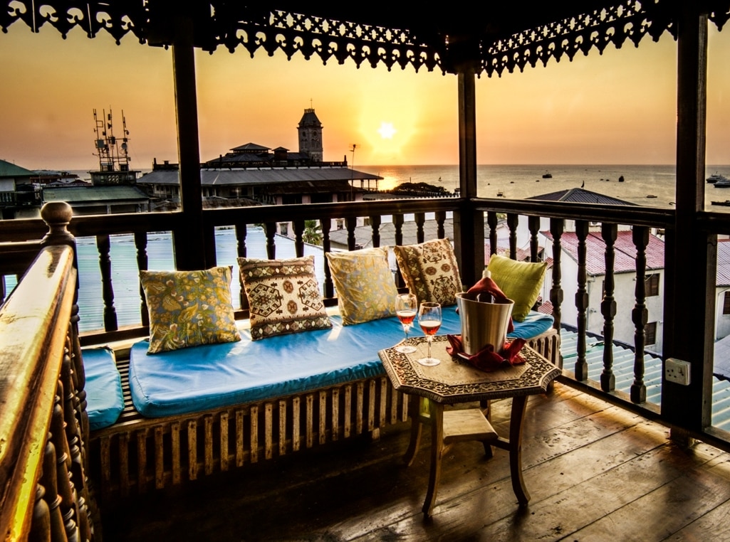 A cozy rooftop veranda features comfortable seating with colorful cushions overlooking the sea. A small table is set with an ice bucket and glasses, while the sunset casts a warm glow across the horizon. The scene captures the tranquil evening atmosphere.