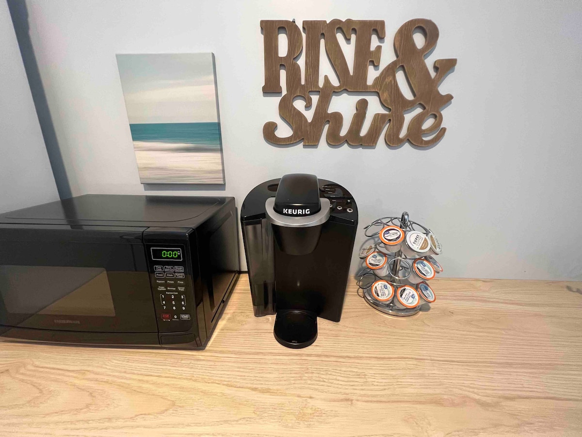 A countertop features a modern microwave alongside a Keurig coffee maker. A circular container holds assorted coffee pods. Above, a wooden sign reading 'Rise & Shine' adds a cheerful touch, complemented by a small artwork depicting a serene seascape.