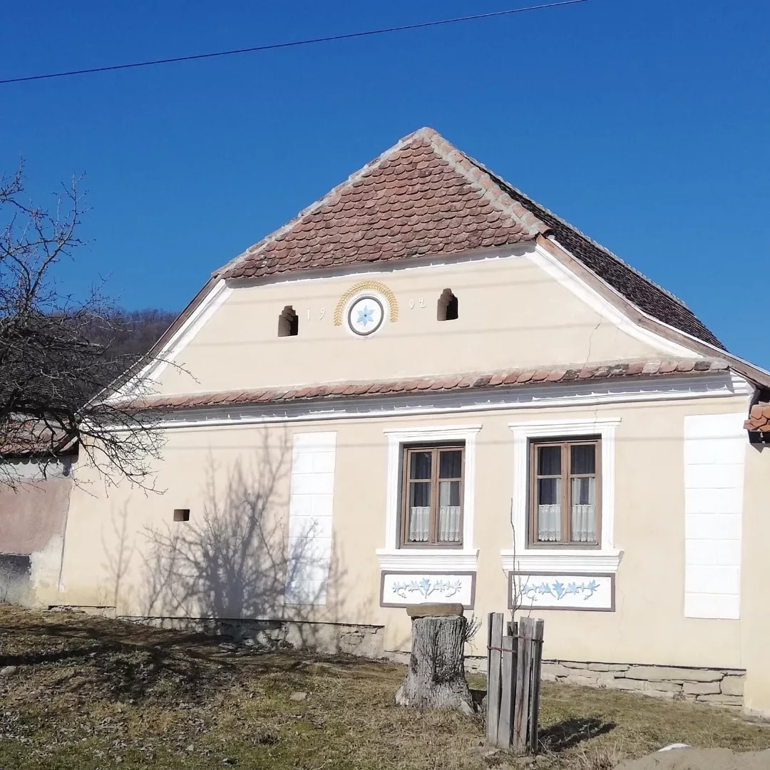 A traditional house displays a gabled roof with terracotta tiles and an ornamental design above the front entrance. Two wooden windows, framed with white shutters, allow natural light to enter. Surrounding greenery and a simple wooden post contribute to the tranquil outdoor setting.