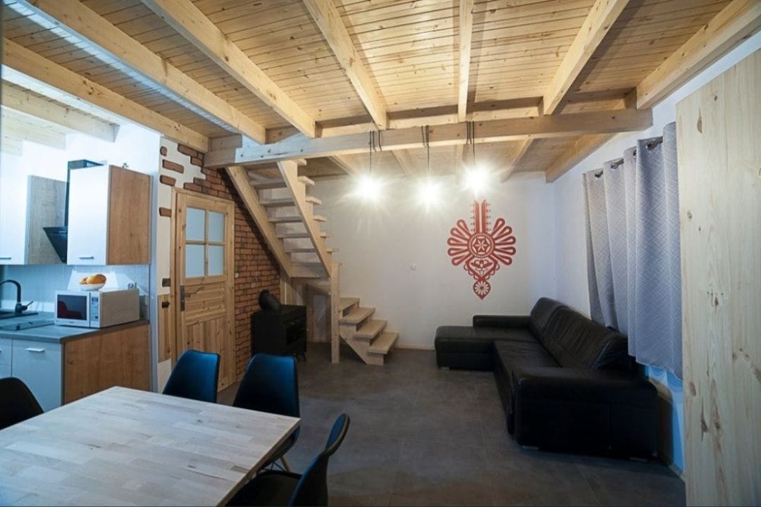 An open living area is illuminated by three pendant lights, featuring wooden beams overhead. A dark leather sofa faces a subtly decorated wall, while a light wooden dining table is positioned nearby. The kitchen is equipped with modern appliances, and a staircase leads to the upper level.