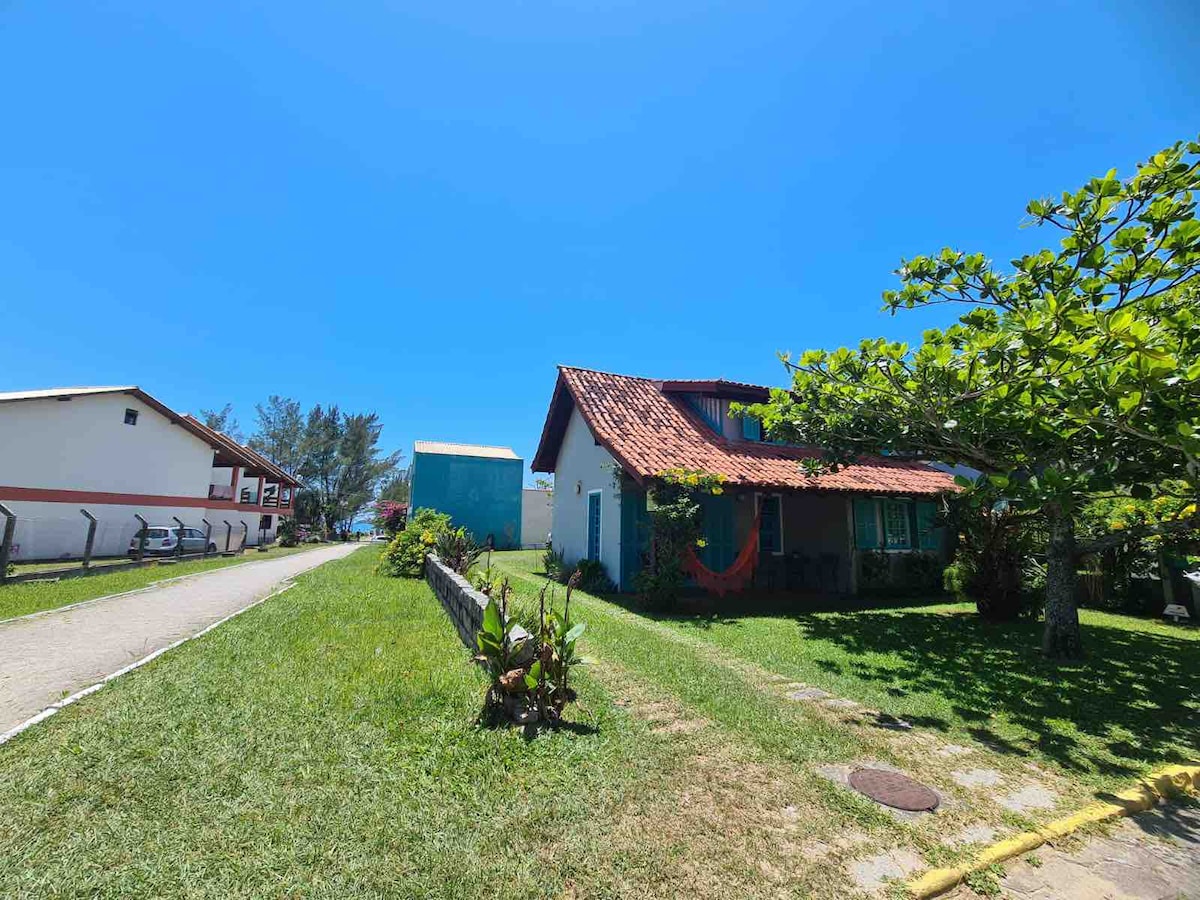 A charming house is set amidst a spacious grassy yard, featuring a red-tiled roof and blue accents. A pathway leads to the entrance, framed by greenery. The surrounding area includes additional buildings, all under a clear blue sky, suggesting a peaceful neighborhood.