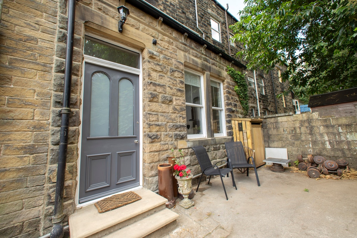 The entrance features a grey door framed by stone walls, with a doormat in front. Two black chairs are positioned beside potted flowers, adding a welcoming touch. A small patio area leads to a natural stone wall, partially obscured by greenery.