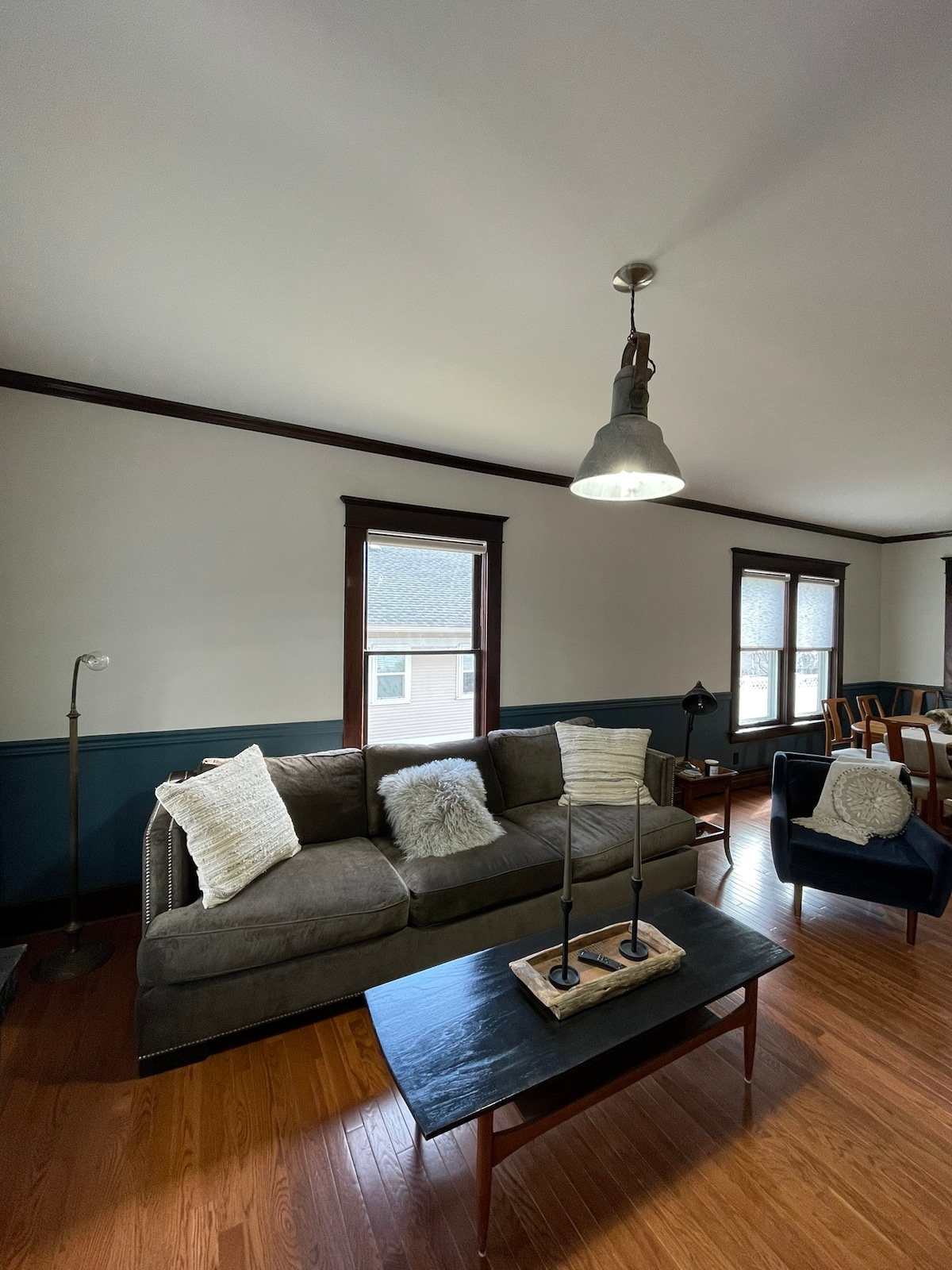A comfortable living area features a dark gray sofa adorned with textured pillows. A wooden coffee table sits in front, holding decorative items. Two large windows allow natural light to fill the space, enhancing the warm wood tones of the flooring and trim.