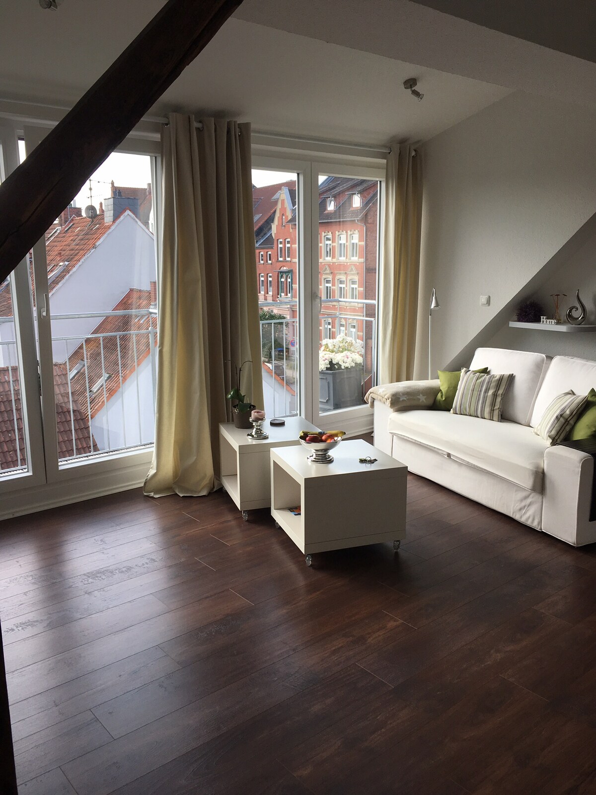 The living area features a white sofa with green accent pillows, complemented by two small coffee tables. Large windows allow ample natural light and provide views of neighboring buildings. Light-colored curtains frame the windows, creating a bright and welcoming atmosphere.