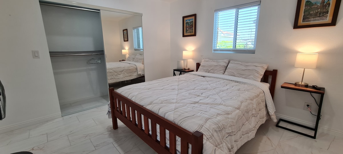 A bright sleeping area features a spacious bed with a neatly arranged white quilt. Two bedside tables with lamps provide soft lighting, while a framed picture adorns the wall. A closet with mirrored doors reflects the light from the window, enhancing the open feel of the space.