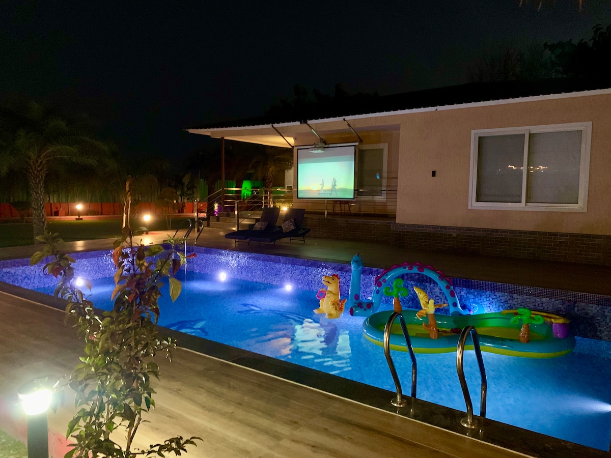 A softly illuminated swimming pool glistens under the night sky, featuring colorful floaties in the water. A screen is positioned nearby, indicating a space for outdoor entertainment. Lush greenery surrounds the area, complemented by ambient lighting on the poolside.