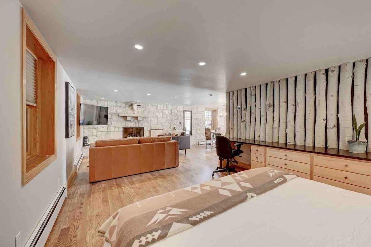 A spacious guest suite features a comfortable king bed with a soft blanket. The room is illuminated by recessed lighting and showcases a cozy seating area with a fireplace. The walls are adorned with a natural wood design, complementing the stone fireplace. A wooden desk is also present.