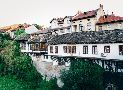 A charming multi-story building is observed with traditional Bulgarian architecture, featuring wooden balconies and a gabled roof covered in shingles. Lush greenery surrounds the base, contrasting with the white and dark wood exterior of the structure. A hillside can be seen in the background, enhancing the natural setting.