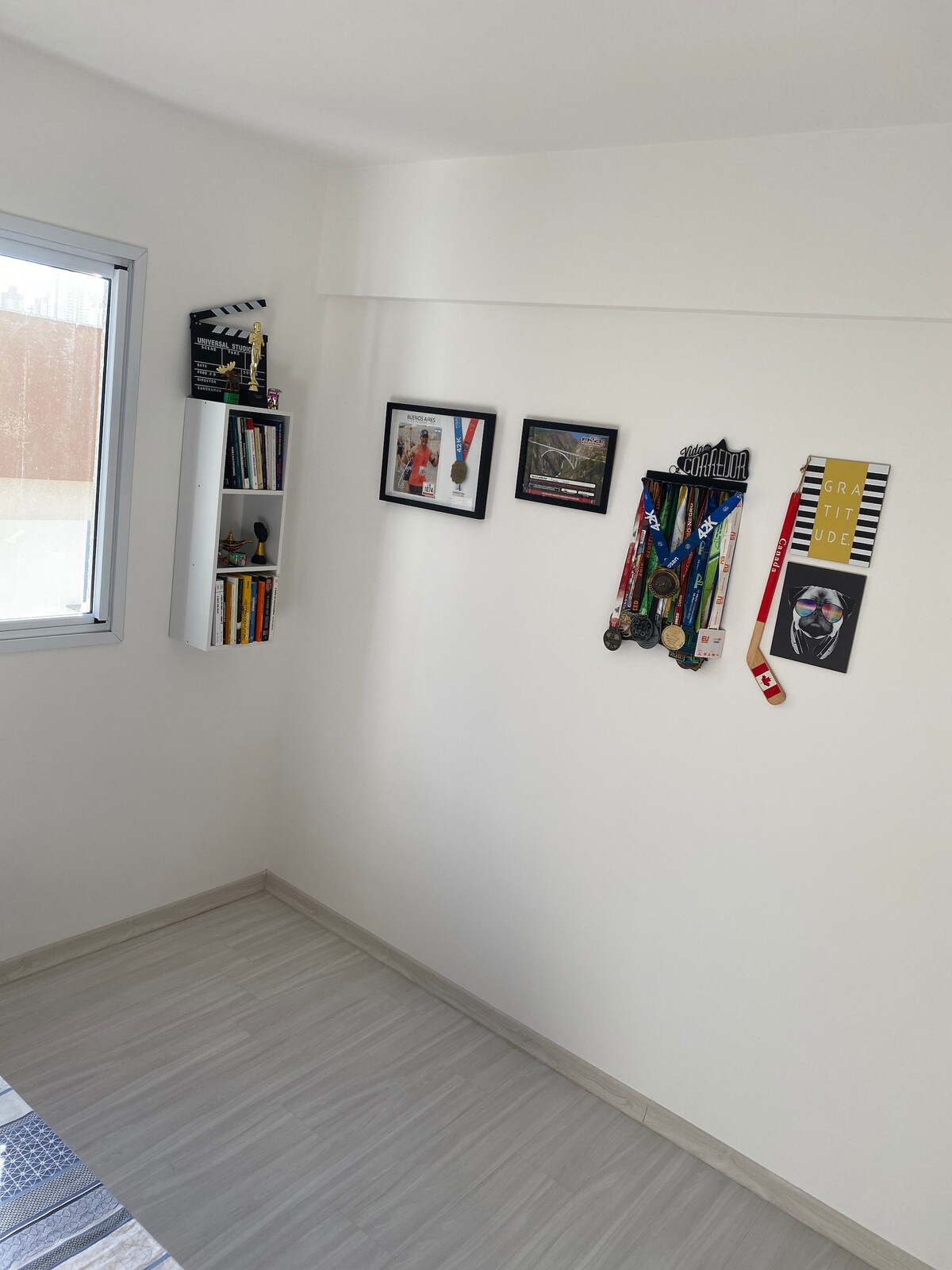 A corner of the room features a small bookshelf stocked with books and framed photographs displayed on the walls. A collection of medals and awards hangs on display, adding character to the neutral-colored space. Natural light filters in through the window.