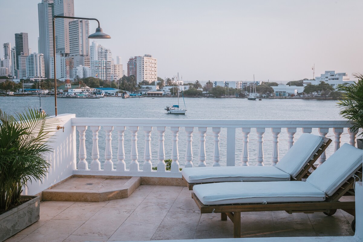 A serene terrace space features two comfortable sunbeds positioned by a railing that overlooks a calm body of water. The distant city skyline is visible, highlighting a harmonious blend of urban and natural scenery. Lush green plants add a touch of nature to the setting.