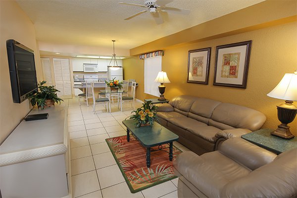 The living area showcases a light-colored leather sofa and an accent table, complemented by warm yellow walls. A flat-screen TV is mounted on a cabinet, while a dining table sits in the background, surrounded by four chairs. Decorative artwork frames the space, enhancing its vibrant atmosphere.