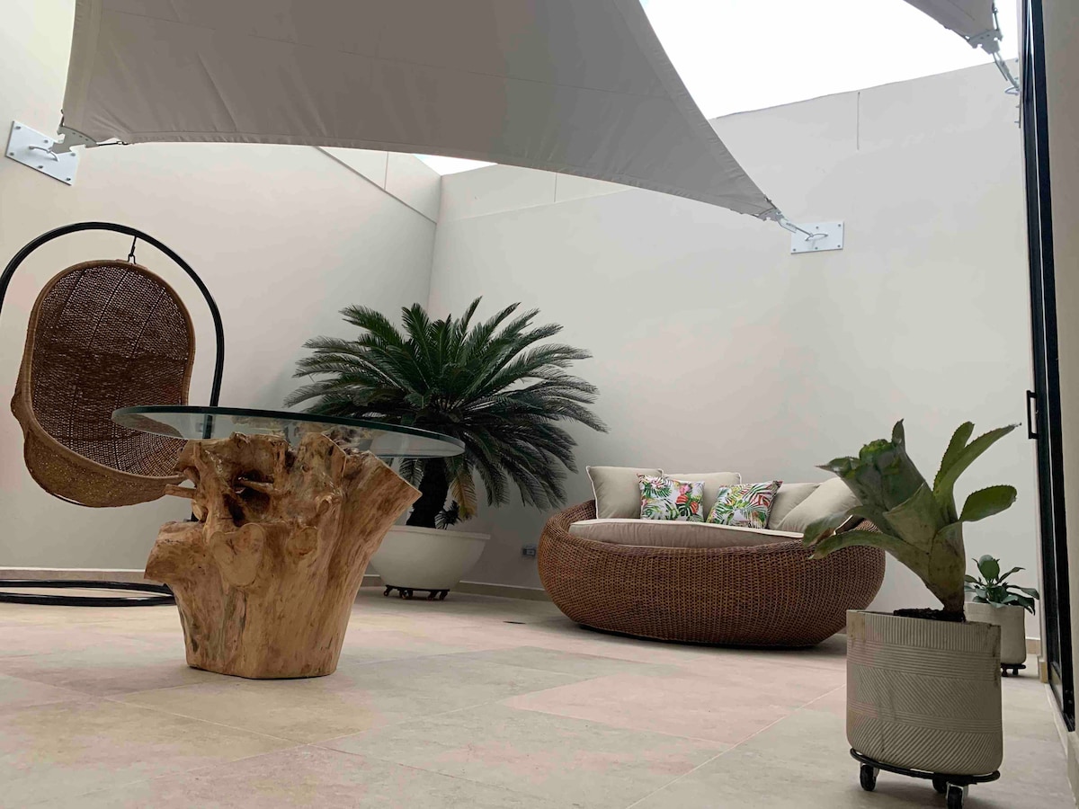 An outdoor terrace is presented with a unique wooden table and a suspended swing chair. Lush green plants are positioned around a comfortable, round seating area adorned with colorful cushions, creating a serene and inviting space for relaxation.