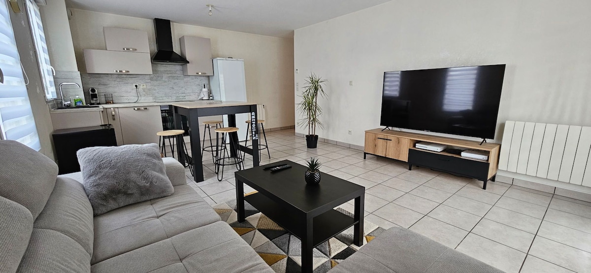 A spacious living area features a light-colored sofa and a coffee table on a patterned area rug. The open kitchen, equipped with modern appliances, is visible along with a rectangular dining table and bar stools. A large television is positioned on a simple media console.