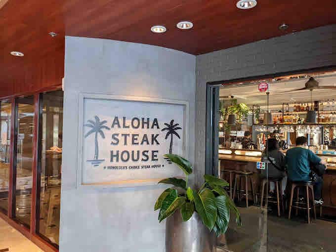The entrance to Aloha Steakhouse is marked by a stylish sign featuring palm trees. Inside, a bar area is visible, with patrons seated at the counter. A potted plant adds a touch of green to the modern decor.