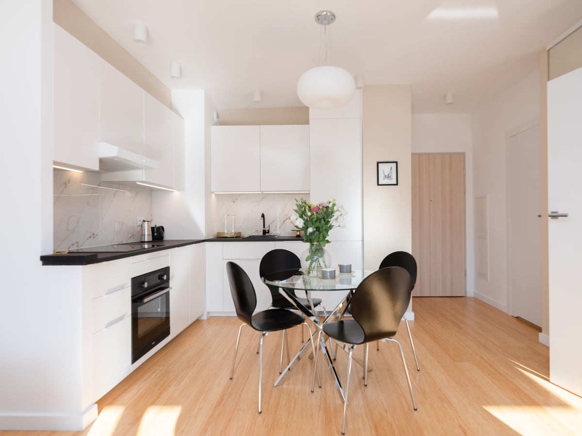 A modern kitchen area features sleek white cabinetry and a marble backsplash. A glass dining table with four black chairs sits in the center, complemented by a vase of flowers. Natural light fills the space from nearby windows, enhancing the clean, minimalist aesthetic.