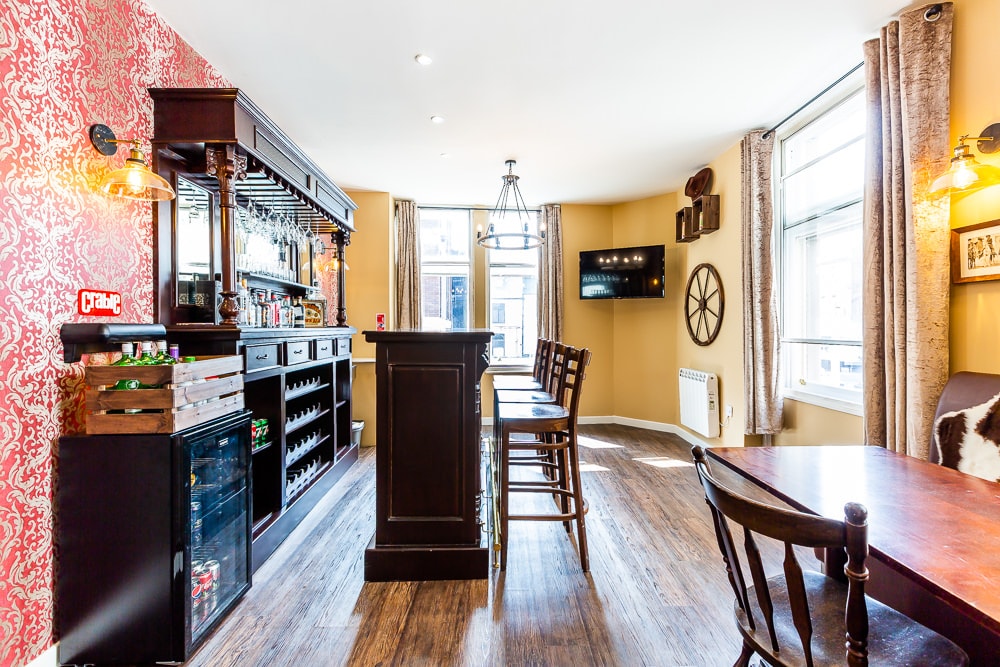 A spacious bar area is highlighted by dark wood cabinetry and multiple seating options. A self-serve bar is stocked with various beverages, complementing the warm tones of the walls. Natural light enters through the large windows, creating an inviting atmosphere for gatherings.