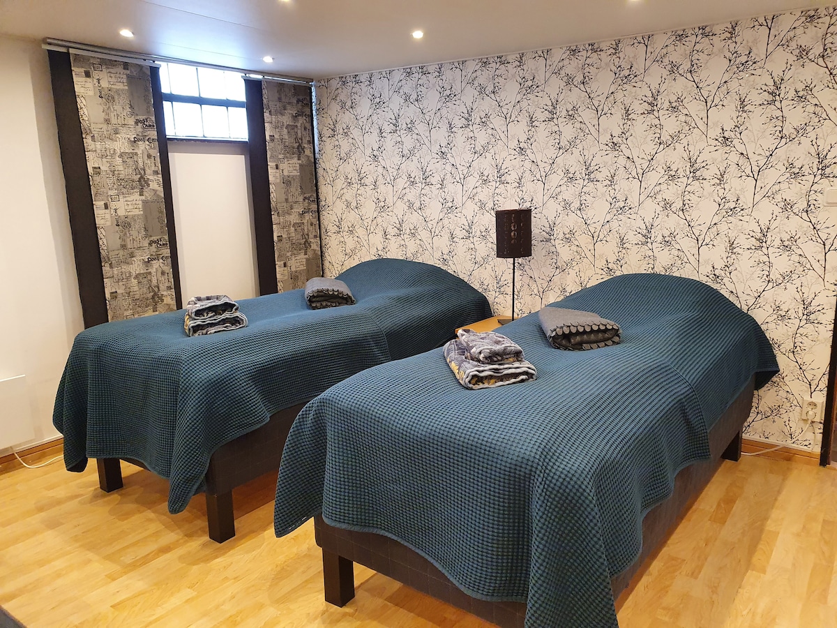The sleeping area features two spacious beds, each covered with textured blue blankets. Coordinated bed linens are neatly arranged at the foot of both beds. A low lamp provides soft lighting, and the walls are decorated with a patterned wallpaper contrasting the wooden floor.