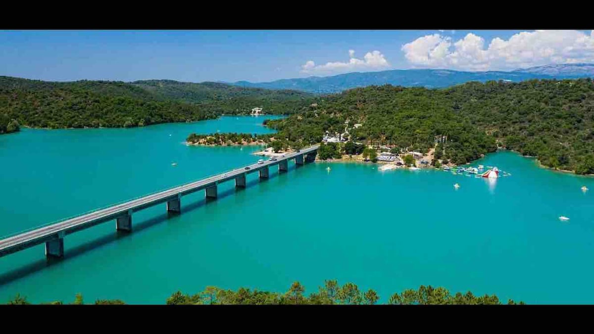 The image captures a scenic view of a vibrant turquoise lake, bordered by lush green trees. A bridge spans across the body of water, connecting both sides. The landscape includes small boats and recreational areas along the shoreline, with hills visible in the background under a clear sky.