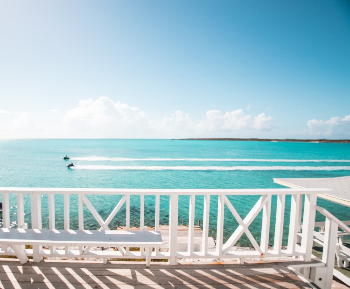 A bright view of the turquoise waters is framed by a white wooden railing. Two jet skis are seen gliding across the water, and the sky is clear with a few fluffy clouds. The serene coastal atmosphere is showcased in this vibrant seaside setting.