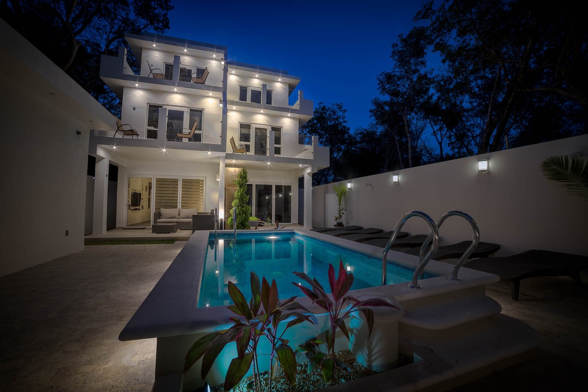 The exterior of the home is illuminated at night, highlighting a tranquil pool bordered by lush foliage. The multi-level structure features balconies with lounge chairs, offering views of the surrounding trees and outdoor space.