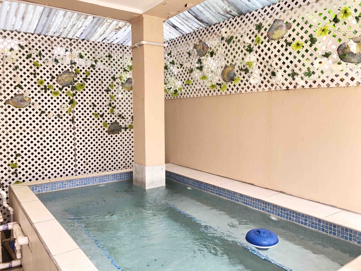 A soaking pool is positioned within a serene area, bordered by a lattice-style wall adorned with green foliage. The clear water reflects natural light, creating a calming environment for relaxation.
