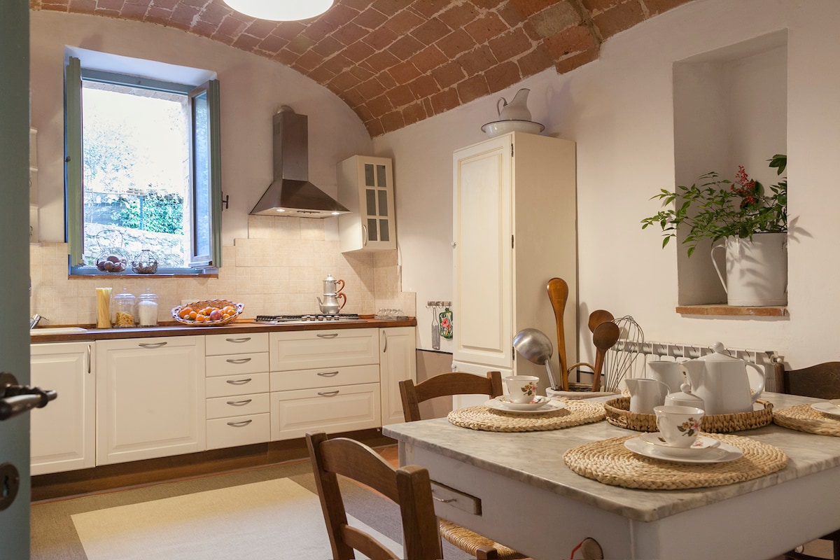 A well-equipped kitchen features arched brick ceilings and a window allowing natural light. Wooden cabinets and a countertop accommodate kitchen appliances and utensils. A dining table, set with plates and cups, is surrounded by wooden chairs, while potted plants add a touch of greenery to the space.