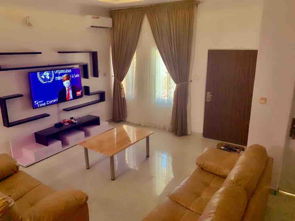 A comfortable living area features two brown leather sofas facing a sleek wall-mounted television. Natural light enters through sheer curtains covering a window, illuminating the glossy, tiled floor. A glass coffee table sits in the center, enhancing the modern aesthetic of the space.