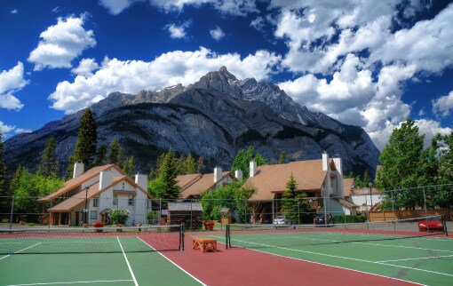 The image showcases outdoor tennis courts framed by lush greenery and framed against the backdrop of a prominent mountain range under a partly cloudy sky. Nearby buildings exhibit a rustic architectural style, adding to the serene, natural setting.