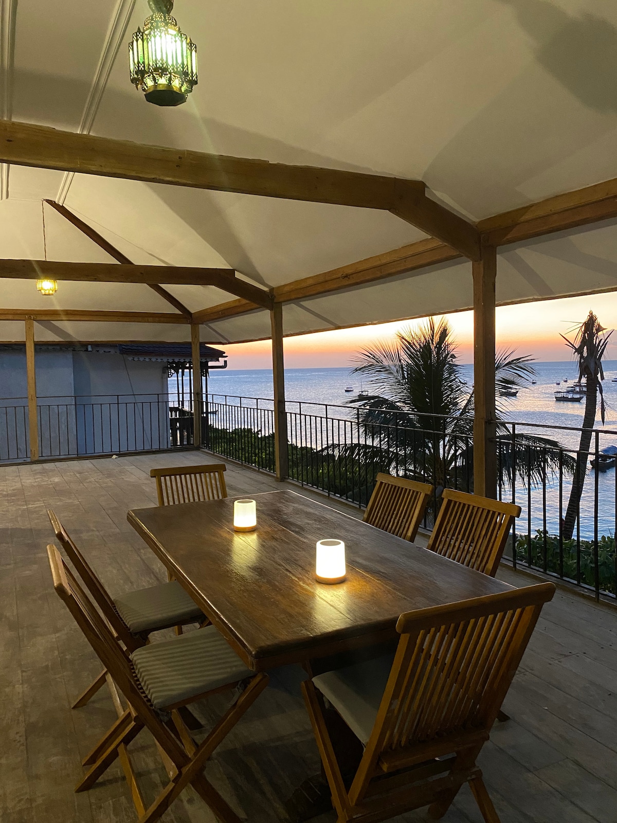 A spacious veranda features a wooden dining table surrounded by six slatted chairs, complemented by gentle lighting from lanterns overhead. The tranquil sea is visible in the background, with a stunning sunset casting soft colors across the horizon.