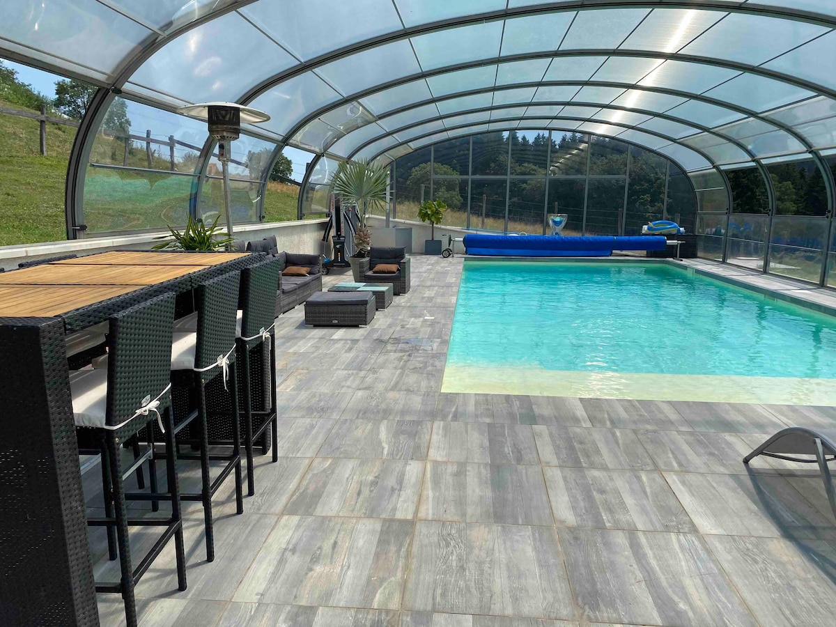 An indoor swimming pool area is seen, enclosed by a transparent roof. Comfortable lounge chairs and a bar counter are positioned along the side, providing a relaxing environment. The pool features clear water, surrounded by a smooth tiled floor, offering a serene setting.