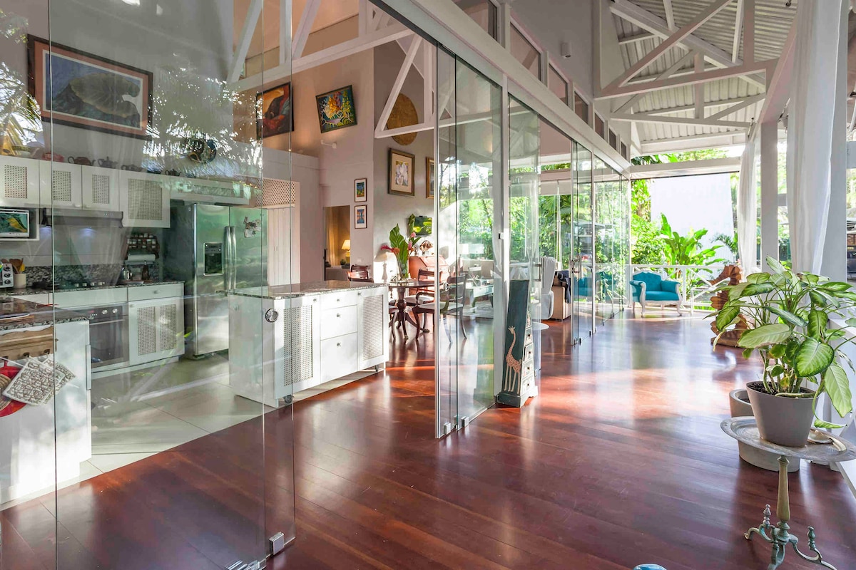 An open layout features a bright interior with wooden flooring and large glass panels that connect the living area to the garden. A fully equipped kitchen can be seen on one side, while dining and lounge areas are illuminated by natural light.