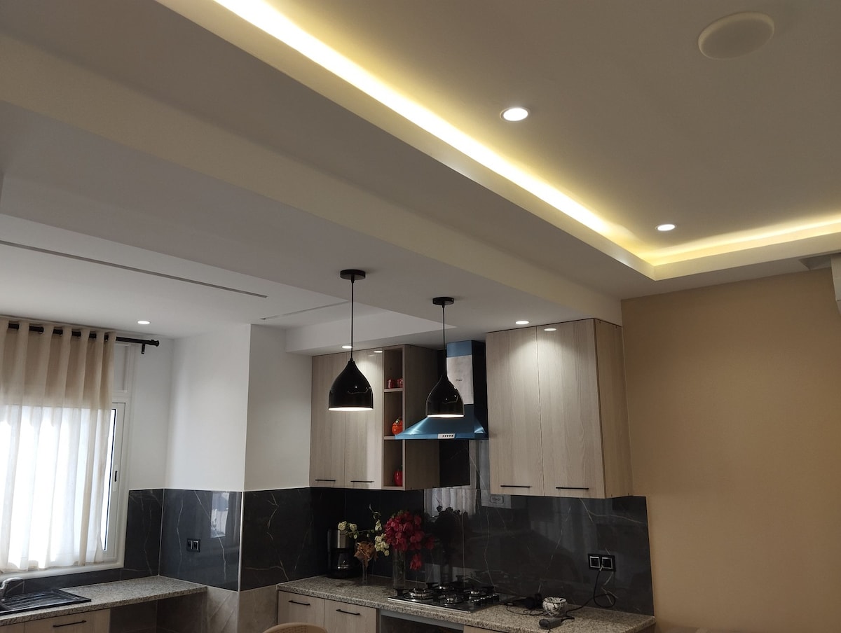 A modern kitchen features sleek cabinetry and a stylish countertop illuminated by pendant lighting. Soft, warm lighting lines the ceiling, enhancing the design. A window allows natural light to fill the space, while decorative elements are placed on shelves and the countertop.