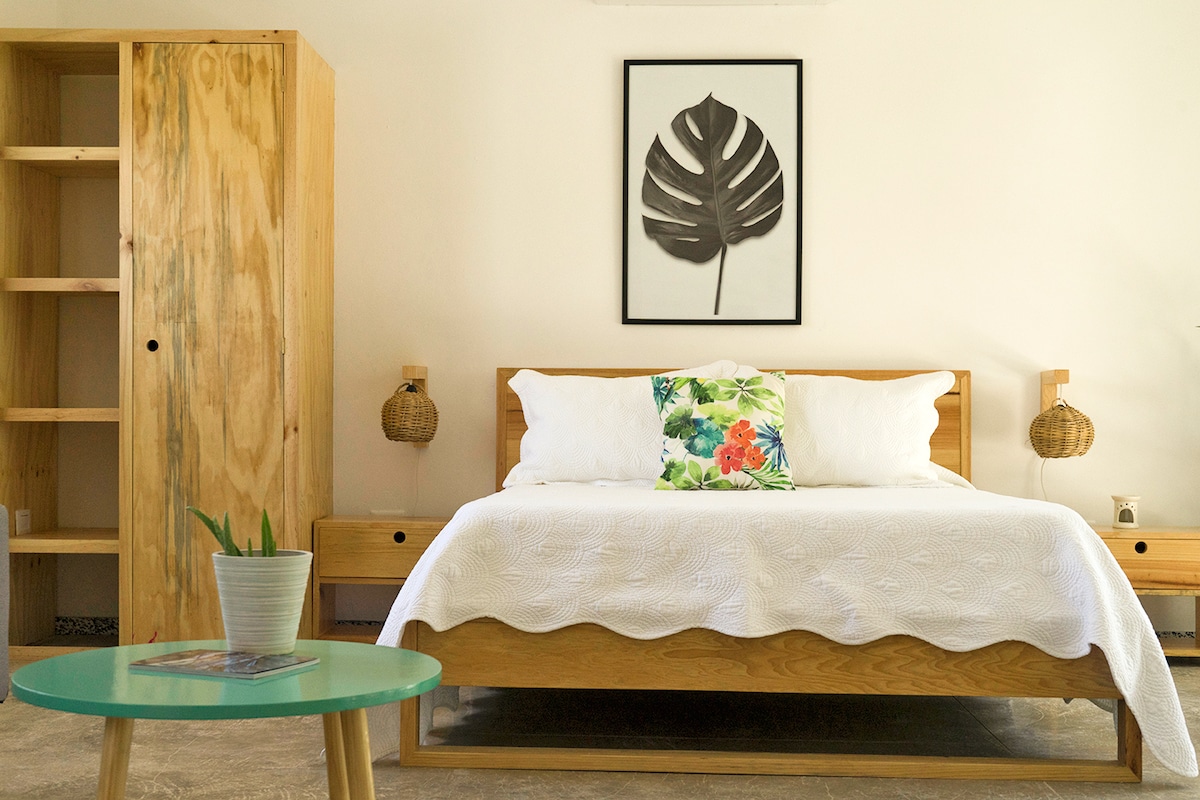 The sleeping area features a wooden bed with a white quilt and decorative pillows, complemented by a matching nightstand. Light wood furniture includes a wardrobe and a round green table with a potted plant, creating a calming and organized space.