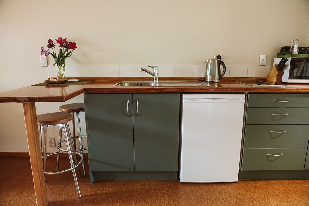 A modern kitchenette features a wooden countertop alongside green cabinetry. Included are a stainless steel sink, a white refrigerator, an electric kettle, and a microwave. Fresh flowers in a vase add a decorative touch to the space. A tall barstool sits beside a small dining area.
