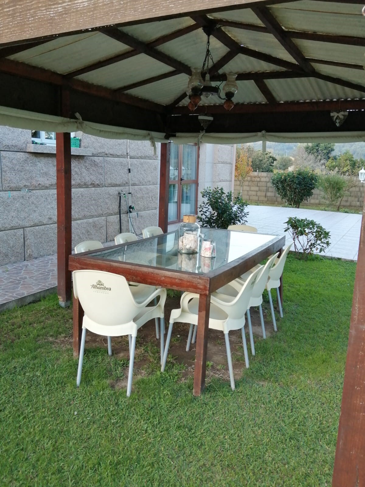 An outdoor dining area features a large glass-topped table surrounded by eight white chairs. The space is shaded by a wooden gazebo with a sloped roof, set on a grassy lawn, and includes a view of landscaped greenery in the background.
