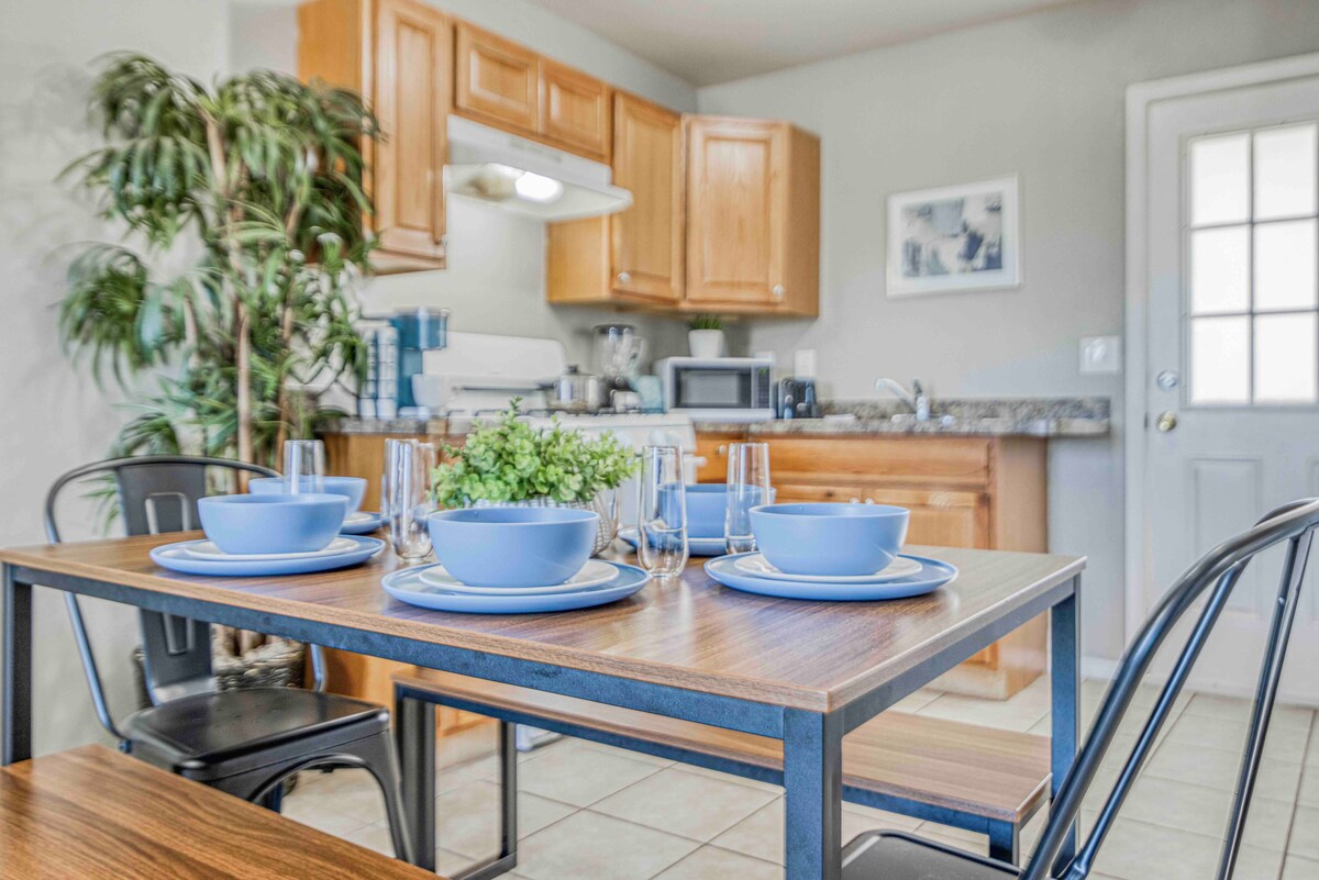 A dining table is set with blue dishes and clear glassware, creating a welcoming space for meals. Behind the table, wooden cabinetry houses kitchen appliances, with a countertop visible. A door provides access to the exterior, while a potted plant adds a touch of greenery.