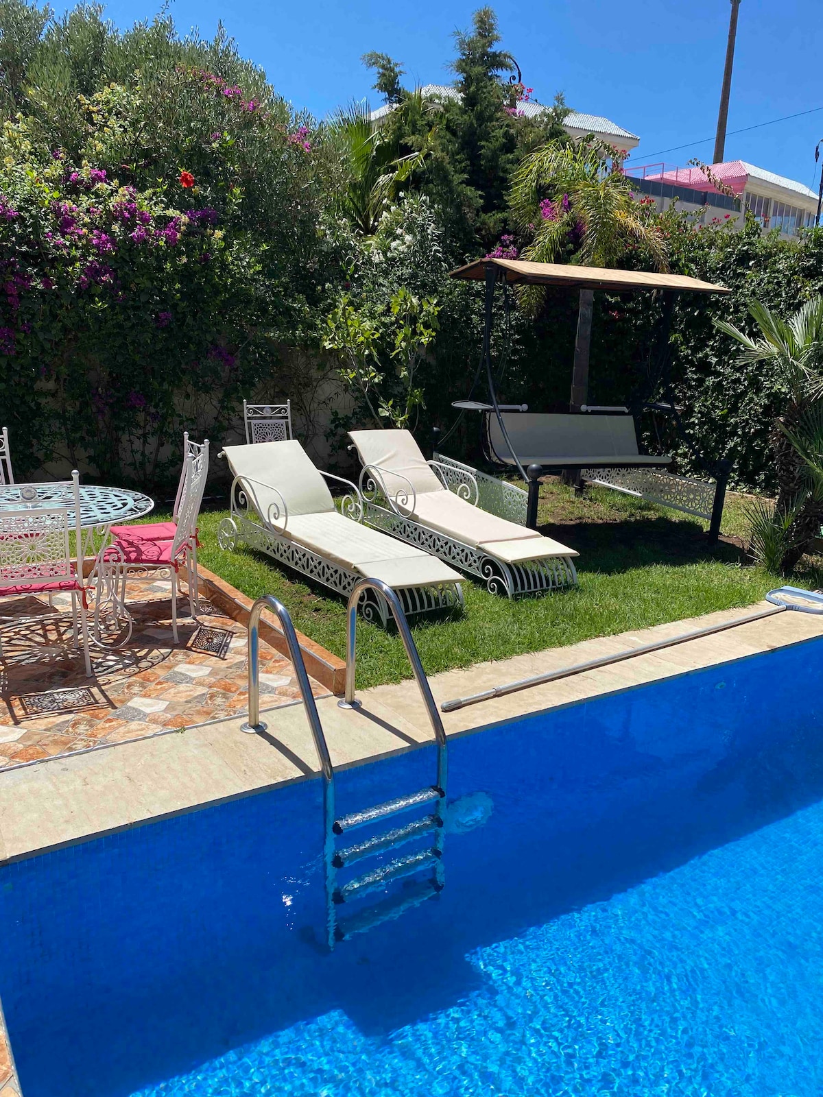 A tranquil outdoor area features a sparkling blue pool with a ladder for easy access. Two white lounge chairs are positioned beside the pool on a grassy section, along with a shaded swing and a round dining table with chairs, surrounded by lush greenery.