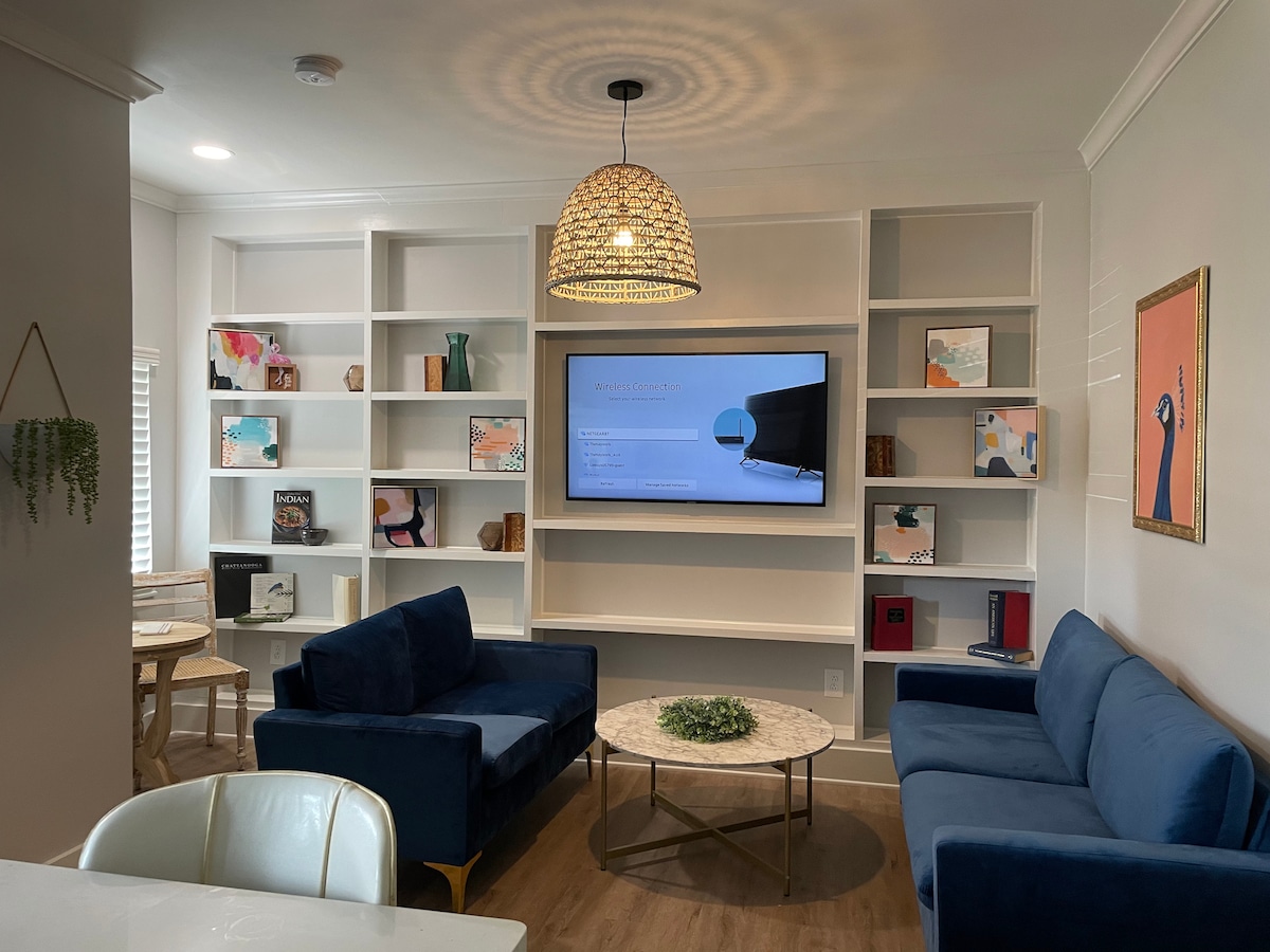 A contemporary living area is presented with navy blue couches arranged around a circular marble coffee table. Wall-mounted shelving displays various decorative objects and books. A large television is set in the center of the shelving, and warm lighting is provided by a pendant lamp overhead.