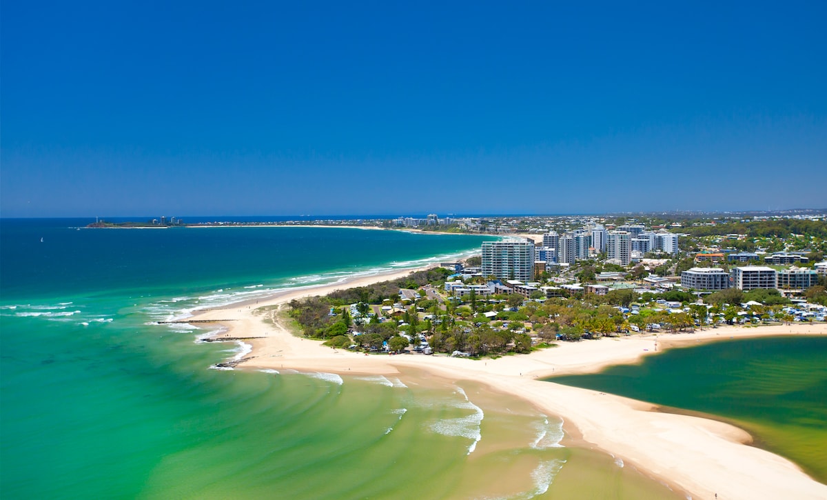 An expansive view captures the coastline of Maroochydore, featuring a blend of sandy beaches and turquoise waters. The shoreline is bordered by residential buildings, with greenery visible in between. The horizon stretches towards a clear blue sky, creating a sense of openness and tranquility.