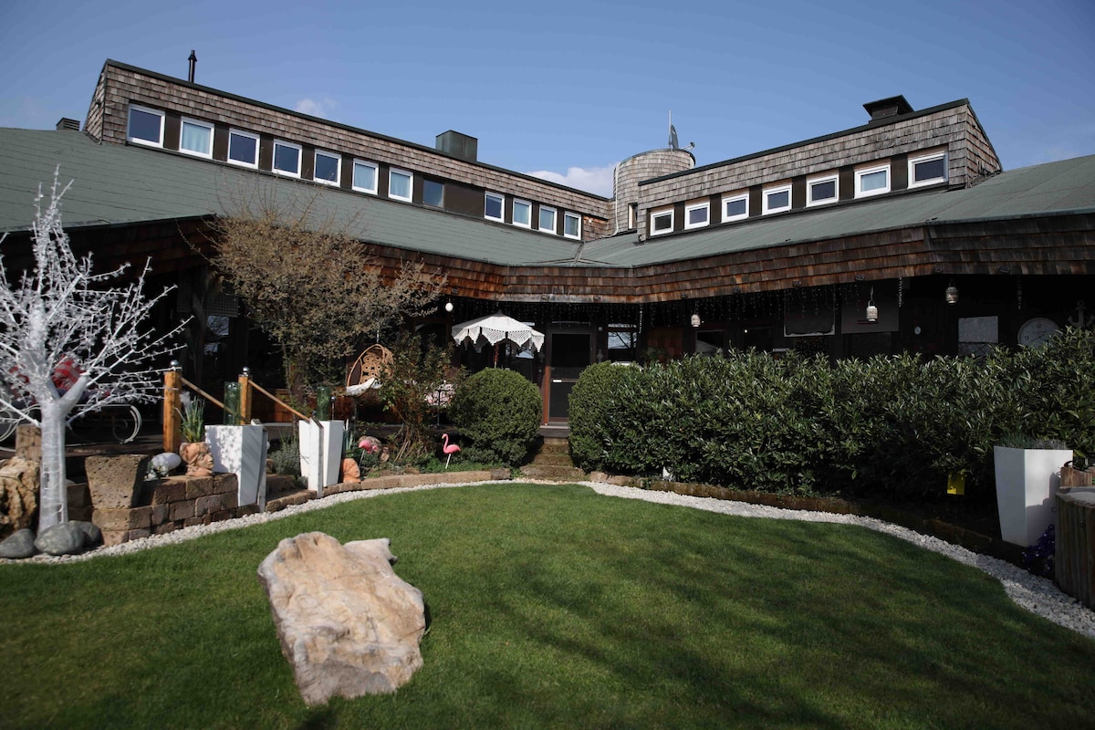 A spacious outdoor area features a well-maintained lawn surrounded by neat hedges. A wooden deck with a table and chairs is visible, accompanied by decorative elements such as a stone sculpture. The building showcases a unique architectural design with multiple roof levels and large windows.