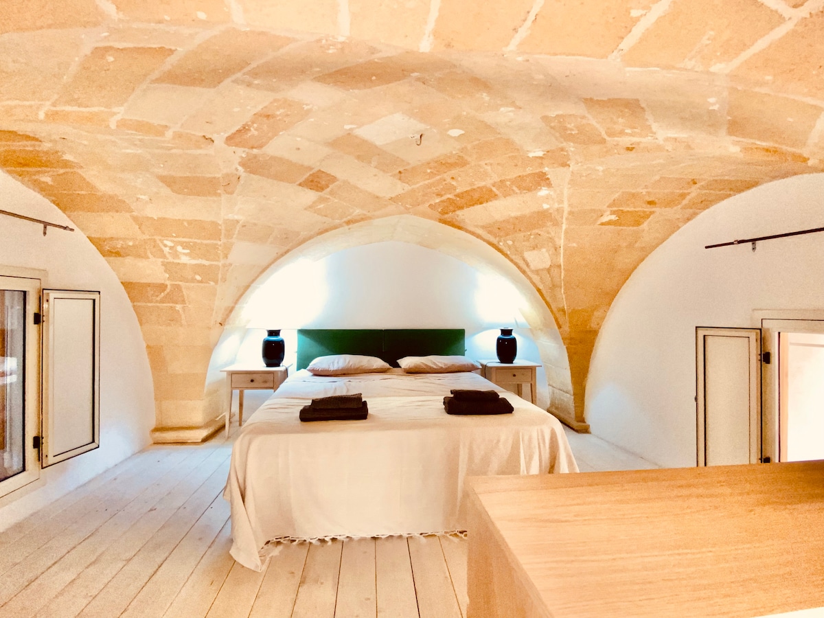 An elevated sleeping area is framed by vaulted ceilings featuring traditional stonework. A spacious bed is complemented by two bedside tables, each adorned with lamps. Natural light enters through small windows, enhancing the airy feel of the room.