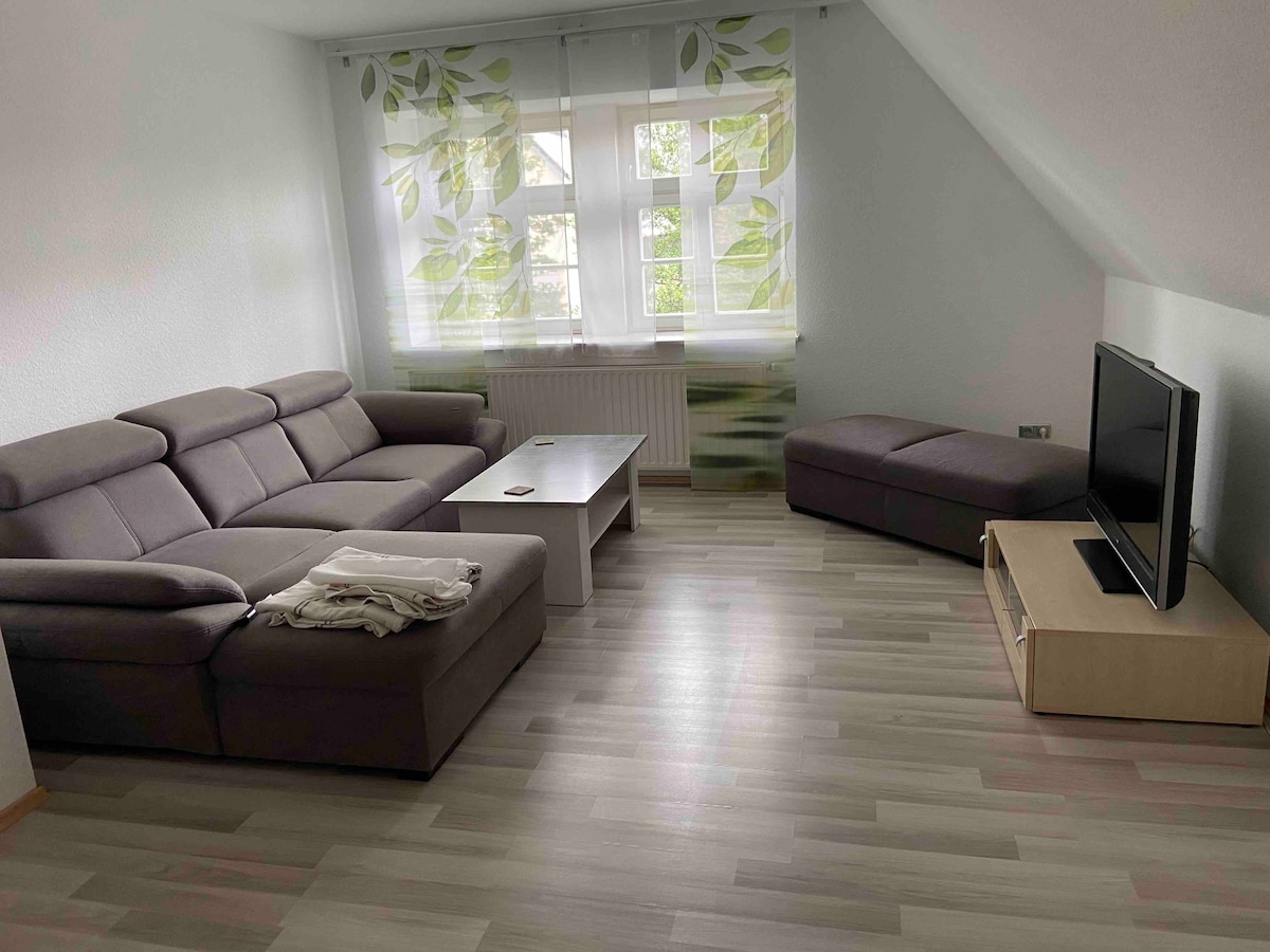 A comfortable living area features a modern sectional sofa in neutral tones, paired with a light wooden coffee table. A flat-screen television is positioned on a media unit. Natural light filters through large windows adorned with sheer leafy curtains, illuminating the pale wooden floor.