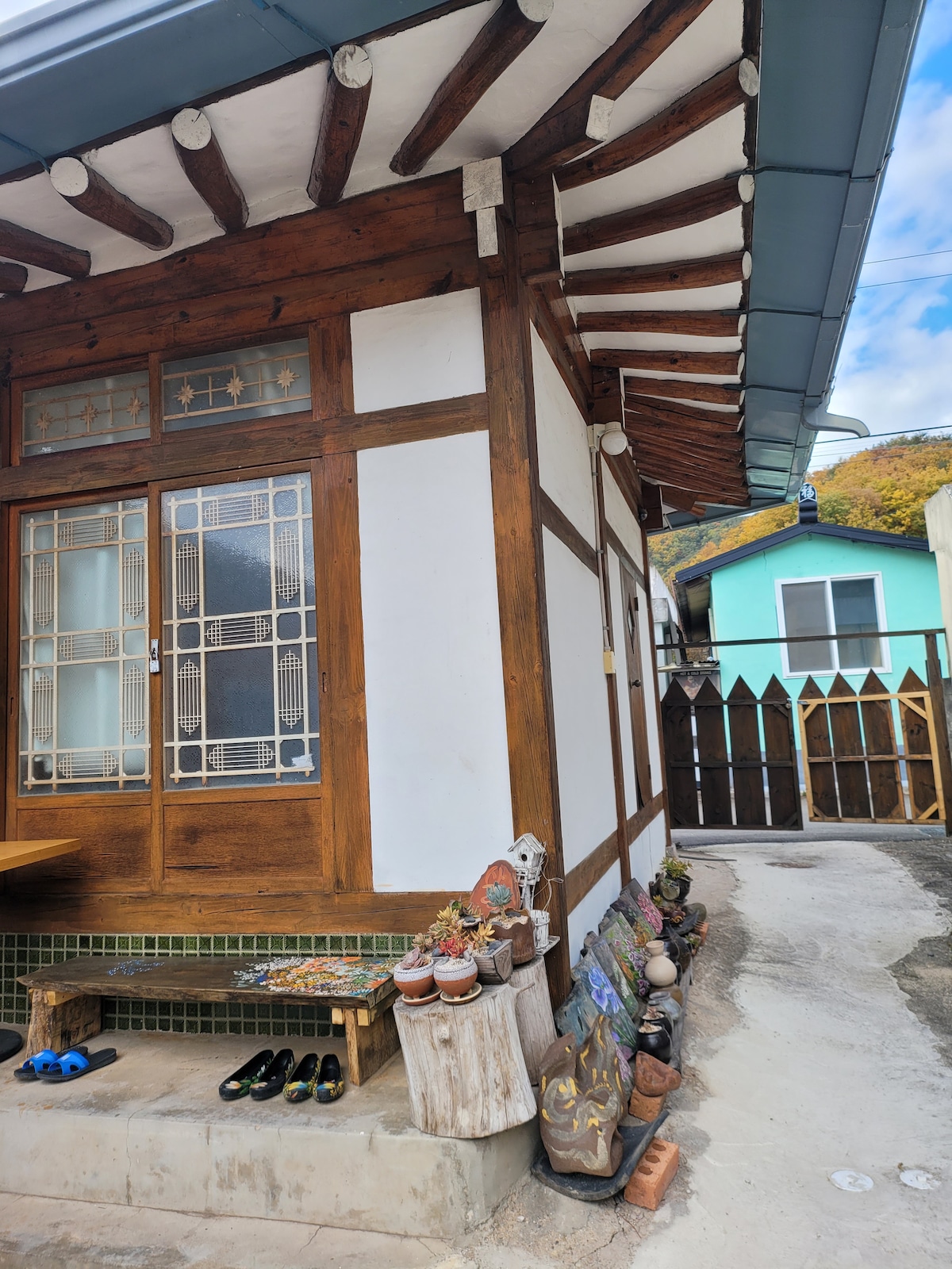 A traditional Korean house is depicted, featuring wooden beams and white walls. A tiled roof is visible above, while a stone bench displays decorative pots along the path. The surrounding area includes a rustic wooden fence, providing a quaint outdoor space.