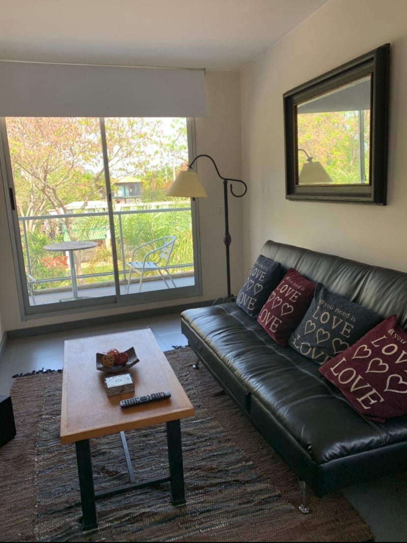 A comfortable living area features a black sofa bed with decorative cushions arranged on it. A wooden coffee table is positioned in front, while a floor lamp and a large mirror are present. Natural light illuminates the space through glass doors to a balcony with a view of greenery.