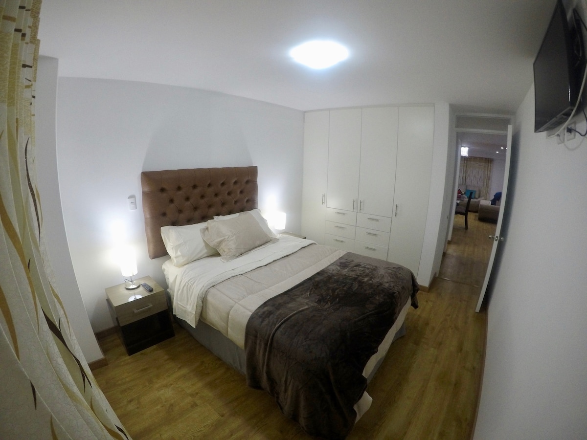 A well-appointed bedroom features a spacious bed with a tufted headboard. Soft bedding complements the warm wood flooring. Nightstands with lamps are positioned on either side, and built-in cabinets provide ample storage. A television is mounted on the wall, adding convenience.