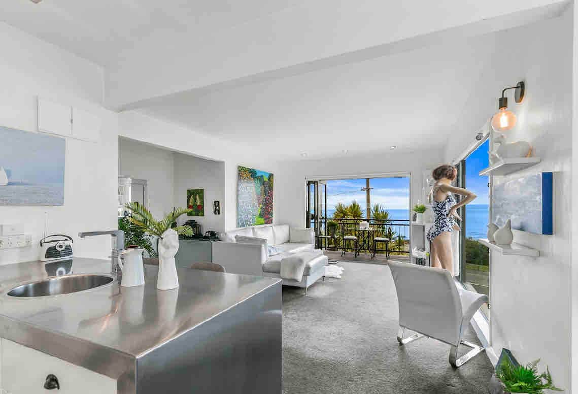 A bright and open living space features sleek modern furnishings, with a spacious seating area adjacent to the kitchen. Large windows offer views of the coastal landscape, while a unique sculpture adds interest to the room. The atmosphere is light and airy.